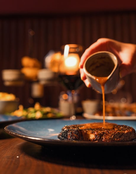 Peppercorn sauce being poured on Steak at Stella Steakhouse with glass of red wine in background